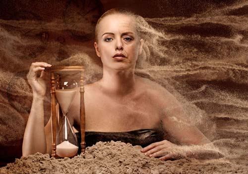 woman in sand up to her chest holding an hourglass, she' changing from young to old